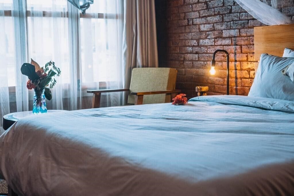 A hotel room with a brick wall, a bed with white linens, a chair, a bedside table with a lamp, and a vase with flowers.