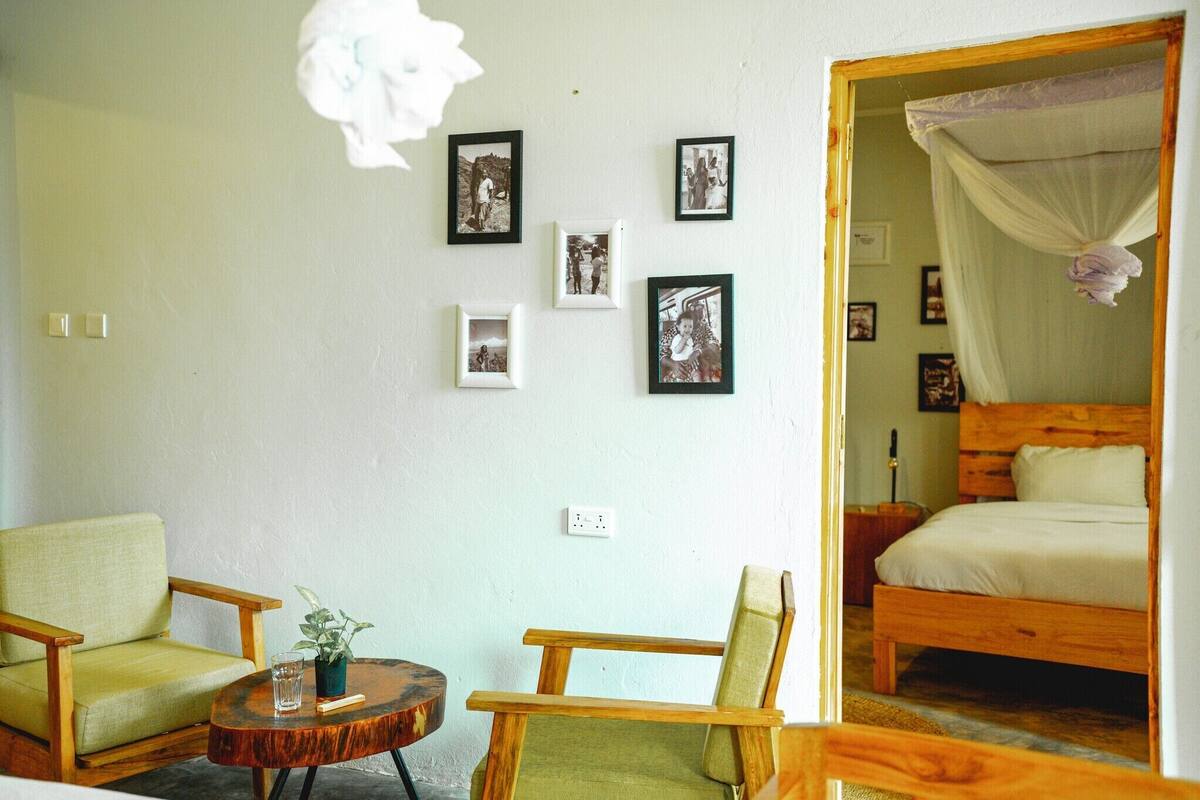 A room with a bed, two chairs, a small table, and a wall with framed pictures.