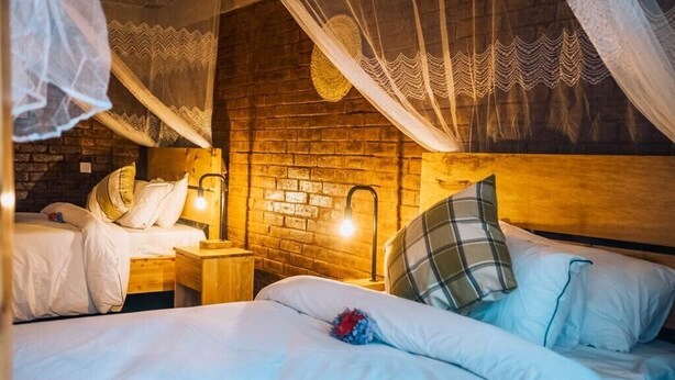 A cozy bedroom with a brick wall, two beds with white bedding, wooden nightstands, and a canopy bed with a decorative hanging. - Rafiki Retreat (Arusha)