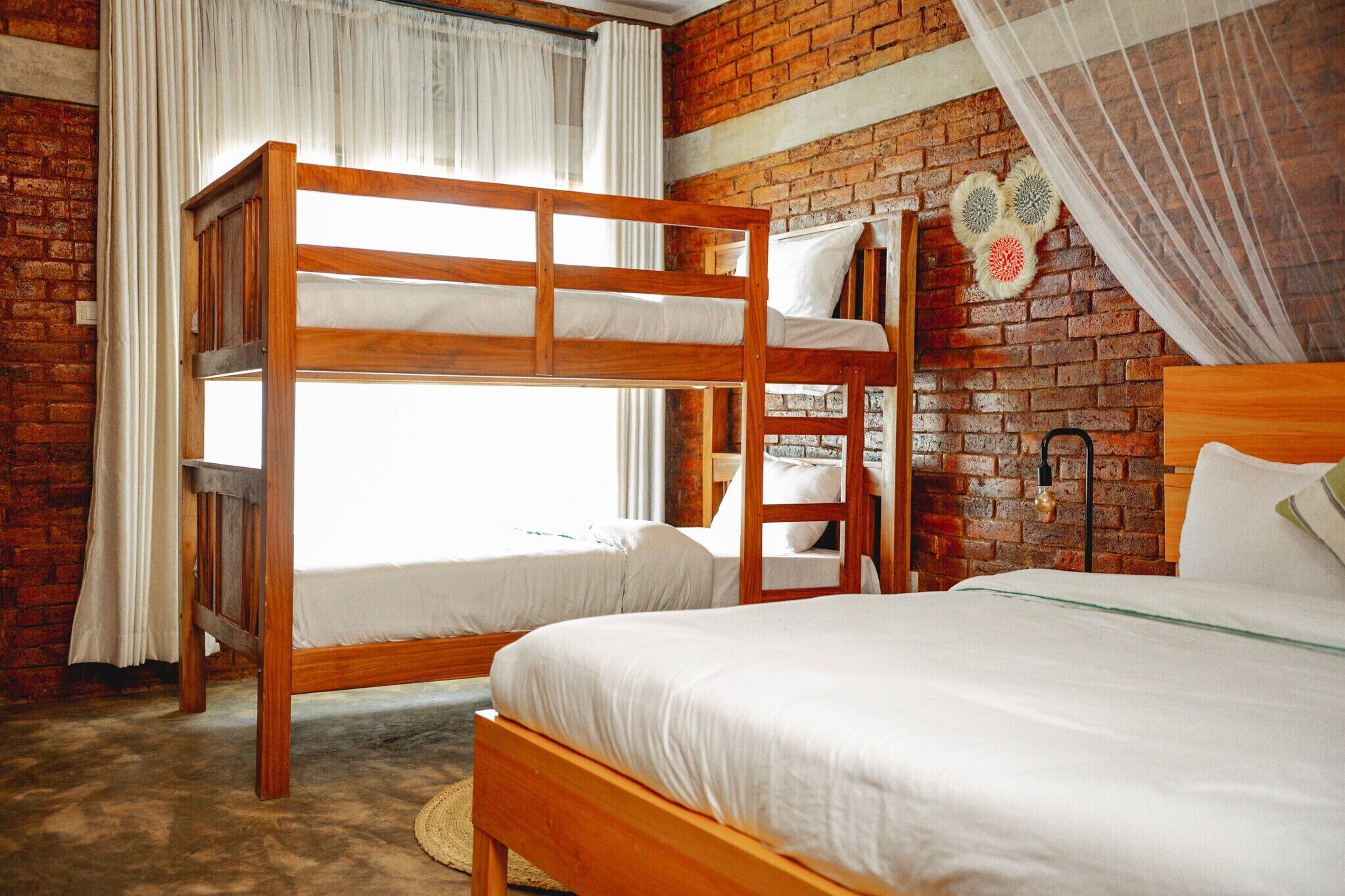 Una habitación con dos literas, una cama individual, una pared de ladrillo, una ventana con cortinas y una lámpara fijada a la pared.
