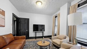 A living room with a brown leather sofa, beige armchairs, a round wooden coffee table, and a flat-screen TV on a stand. - Merion Village Residences 1166 (Columbus)