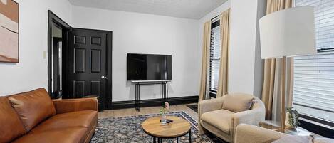 A living room with a brown leather sofa, beige armchairs, a round wooden coffee table, and a flat-screen TV on a stand.