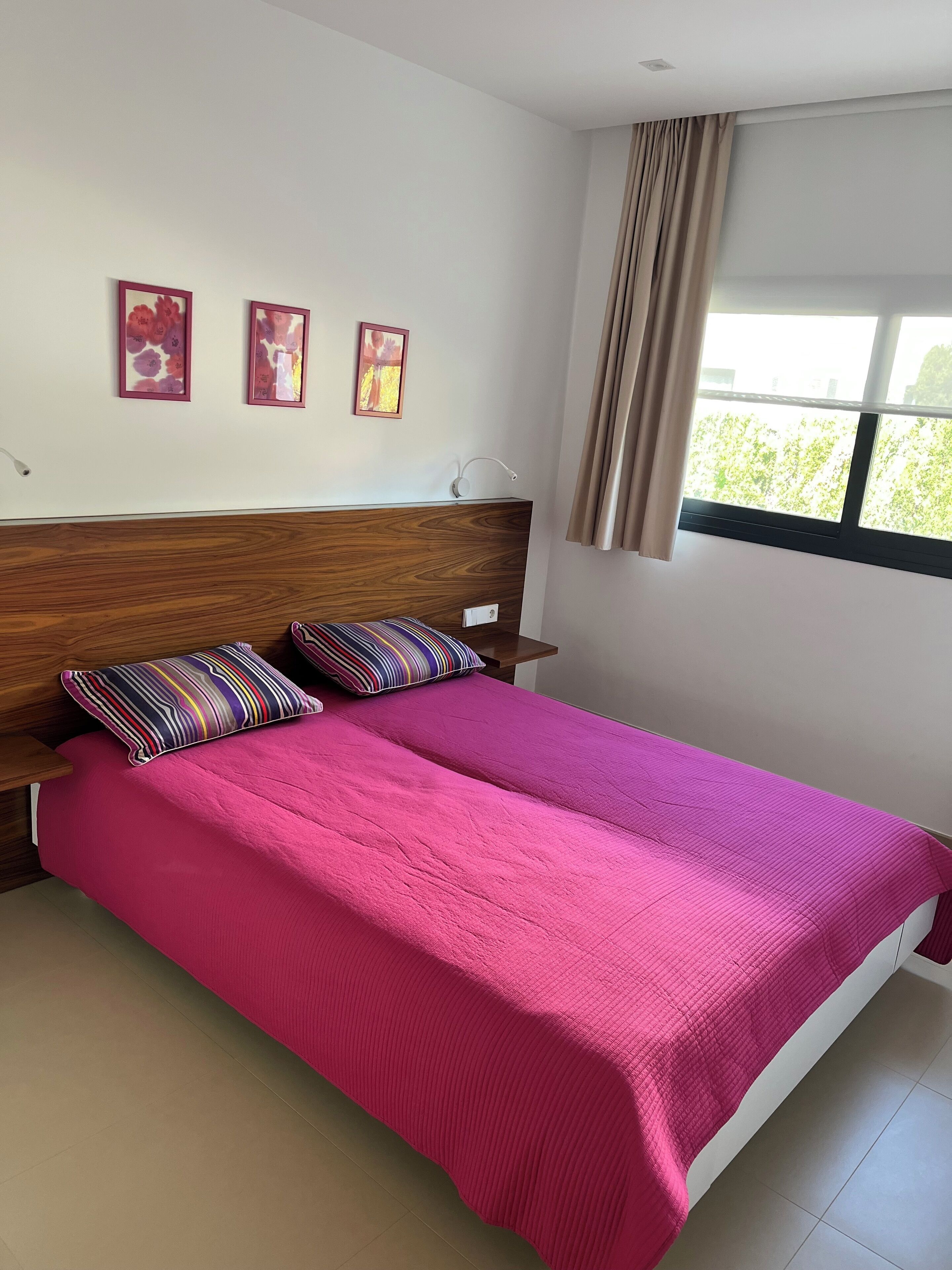 Ein Schlafzimmer mit einem hölzernen Kopfteil, einer rosa Bettdecke und zwei gestreiften Kissen. An der Wand hängen drei gerahmte Bilder und es gibt ein Fenster mit Vorhängen.