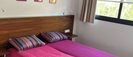 Ein Schlafzimmer mit einem hölzernen Kopfteil, einer rosa Bettdecke und zwei gestreiften Kissen. An der Wand hängen drei gerahmte Bilder und es gibt ein Fenster mit Vorhängen.