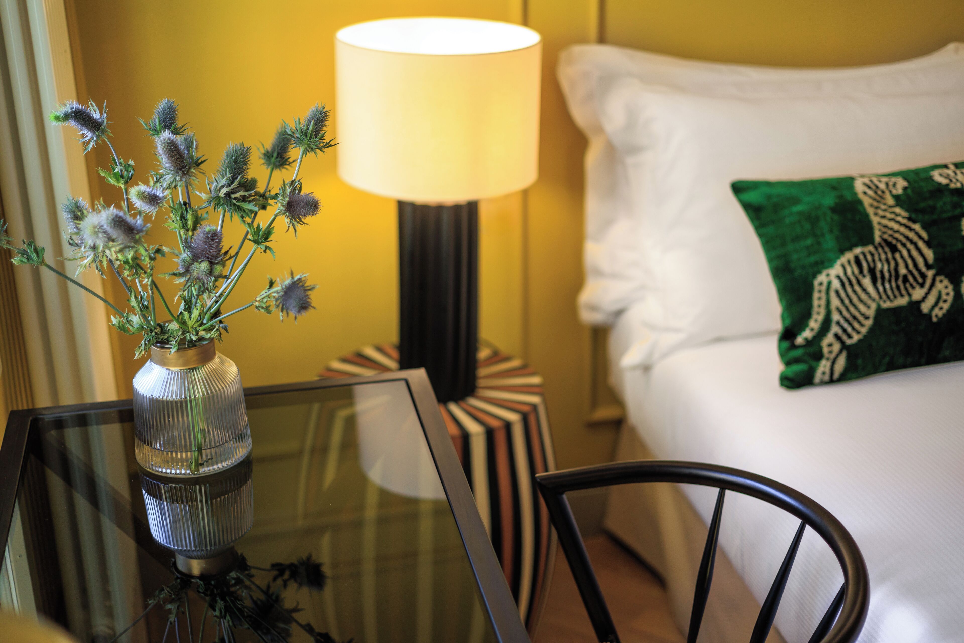 A hotel room with a glass table, a vase with dried flowers, a lamp, a striped side table, a bed with white bedding and a green pillow, and a black chair.