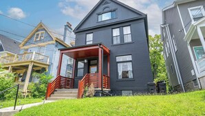 A two-story house with a dark gray exterior, a wooden porch with a red railing, and a visible front door. - Walk to Xavier 5BR 2 Kings Sleeps 10 (Cincinnati)