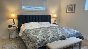 Une chambre à coucher bien rangée, avec un grand lit, deux lampes de chevet, une table de chevet avec une horloge, un banc et un tapis.
