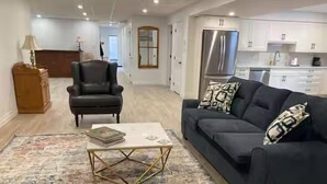 Un salon moderne avec un canapé foncé, une table basse et un tapis à motifs. On y trouve également un meuble en bois et un fauteuil noir. La cuisine, visible à l’arrière-plan, est dotée de meubles blancs et d’appareils électroménagers en acier inoxydable.