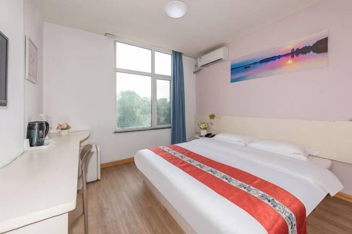 A hotel room with a bed, a desk, a window, and a painting on the wall.