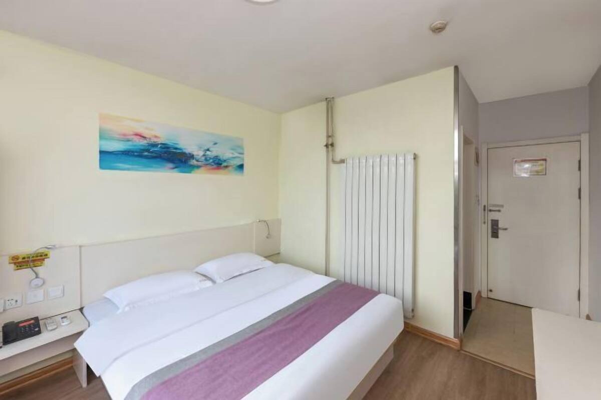 A hotel room with a bed, a bedside table, a radiator, and a door.