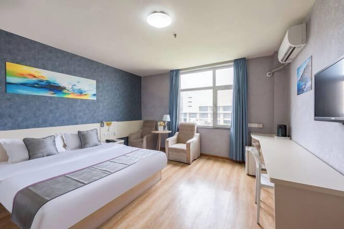 A hotel room with a large bed, a desk, two armchairs, a painting on the wall, and a window with curtains.