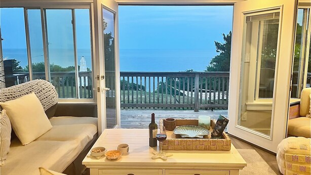 A cozy living room with a sofa, a coffee table with snacks and drinks, and a view of the ocean through large sliding glass doors. - Panoramic Cape Cod Bay Views with Private Sandy Beach (Plymouth)