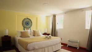 A neatly made bed with a beige blanket and pillows, a decorative wall hanging, a bedside table with a lamp, and a radiator. - Casa Pumagui (Vicuña)
