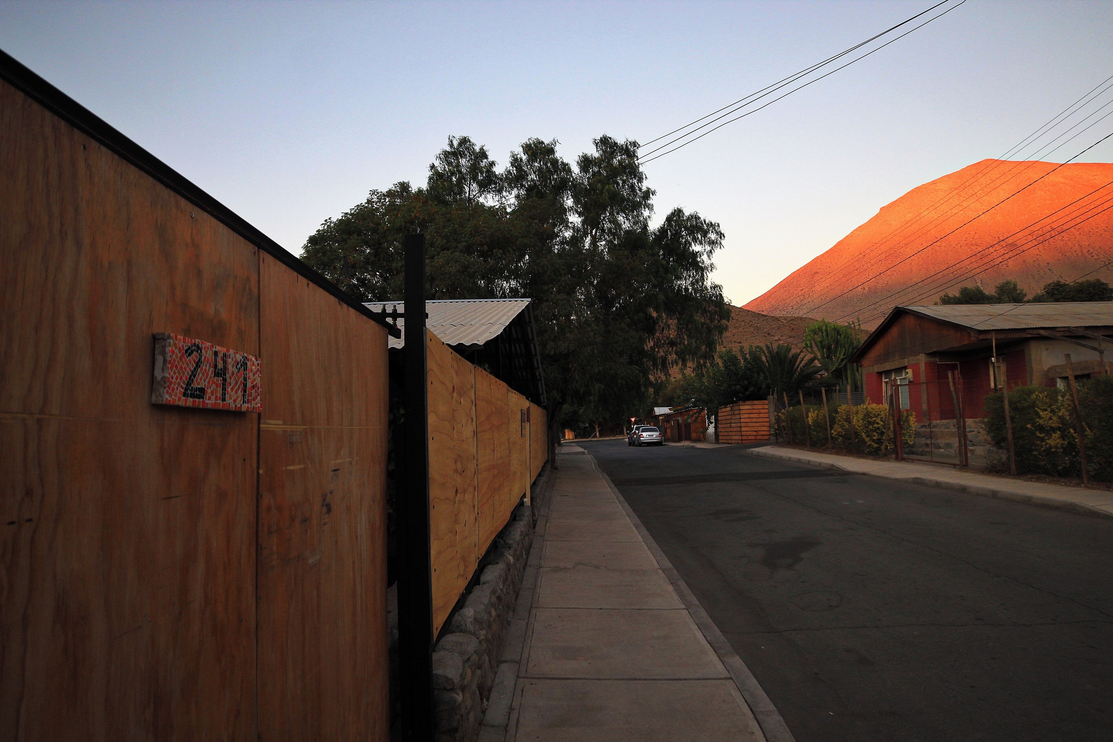 Vista de la calle con una cerca de madera, un letrero con 'ZIM' y un edificio con techo rojo al fondo.