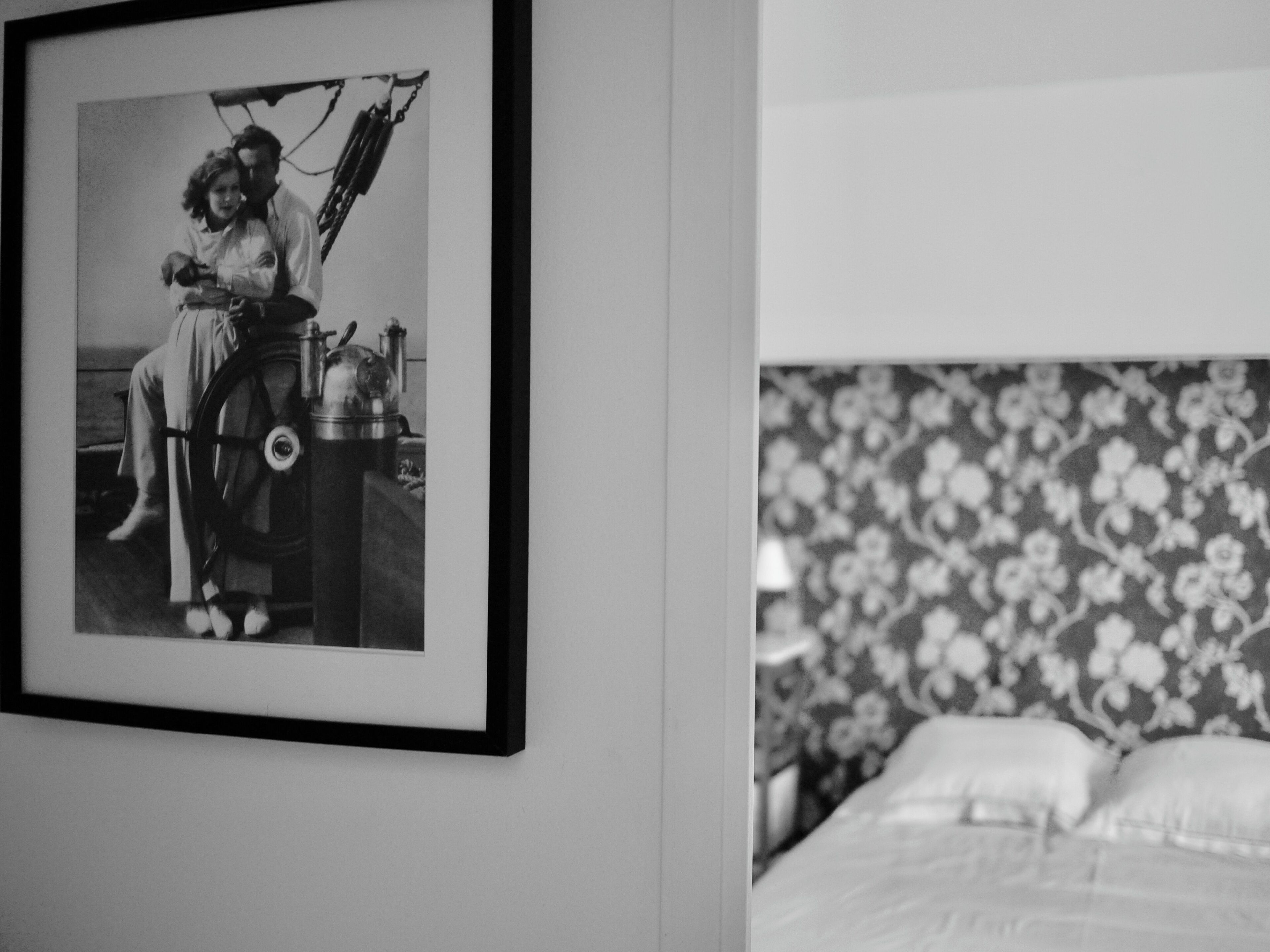 Une photographie en noir et blanc encadrée représentant une personne à vélo, un lit avec une tête de lit à motifs floraux et un mur uni.