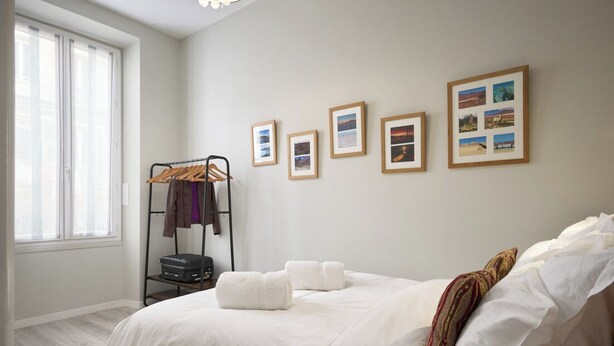 A bedroom with a large bed, a window, a wall-mounted drying rack, and several framed pictures. - Sunlight Properties GISELE: Hyper centre, 1 bedroom with bright modern living (Nice)