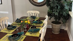 A dining area with a wooden table set for six, surrounded by white chairs. A large mirror hangs on the wall, and a potted plant is to the right.