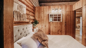 A wooden cabin bedroom with a tufted headboard, a bedside lamp, and a window with a view. - 76gs - Genuine Log Cabin - Wifi - Pets Ok - Sleeps 4 (Deming)