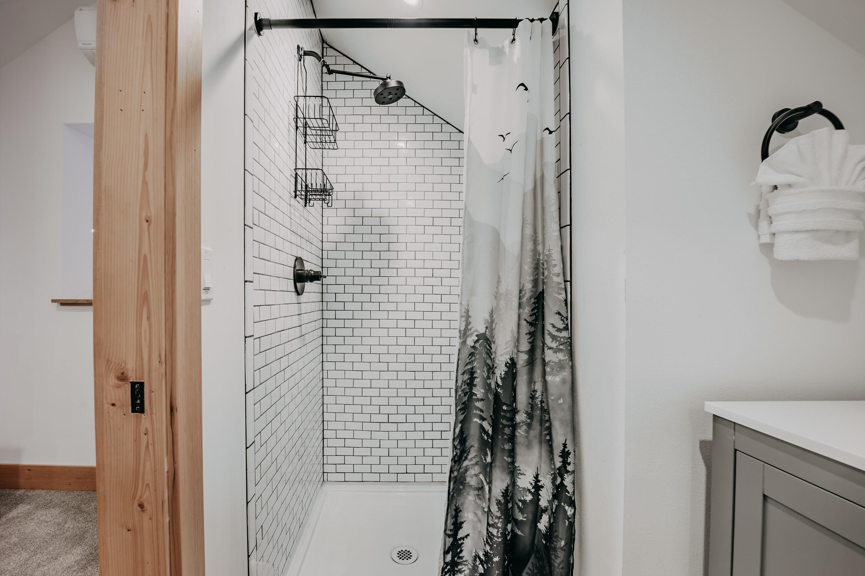 A shower area with a white tiled wall, a showerhead, and a curtain with a forest design.