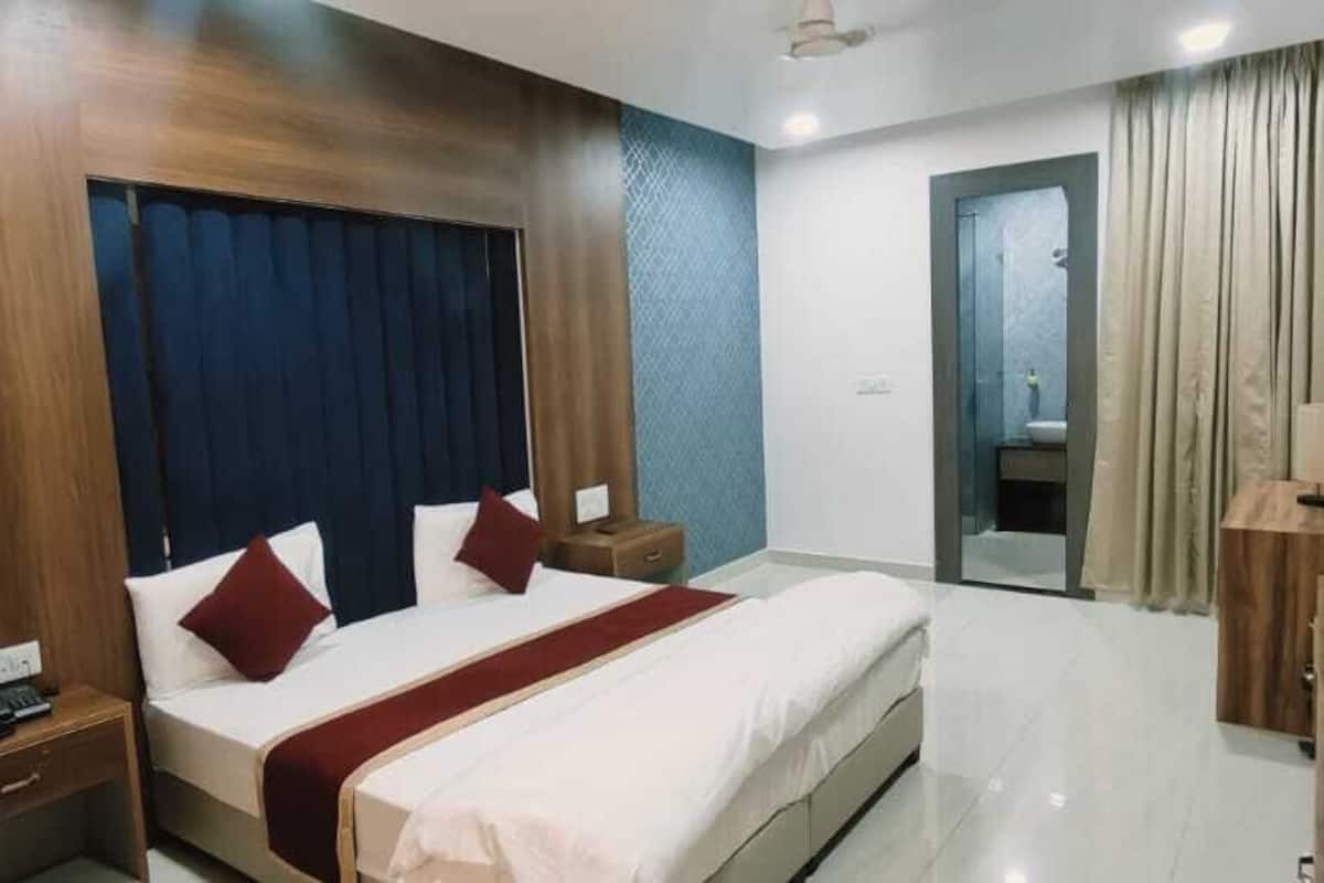 Quarto de hotel com uma cama grande, uma mesa de cabeceira de madeira, televisão e banheiro visível através de uma porta aberta.