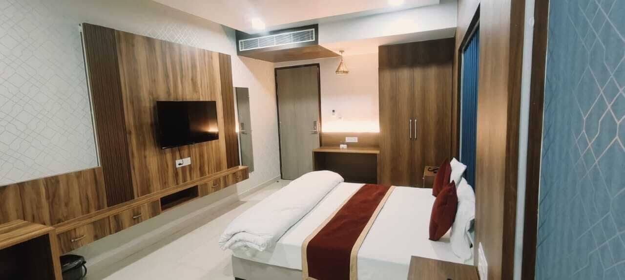 A modern hotel room with a large bed, a flat-screen TV mounted on the wall, a wooden wardrobe, and a small desk with a lamp.