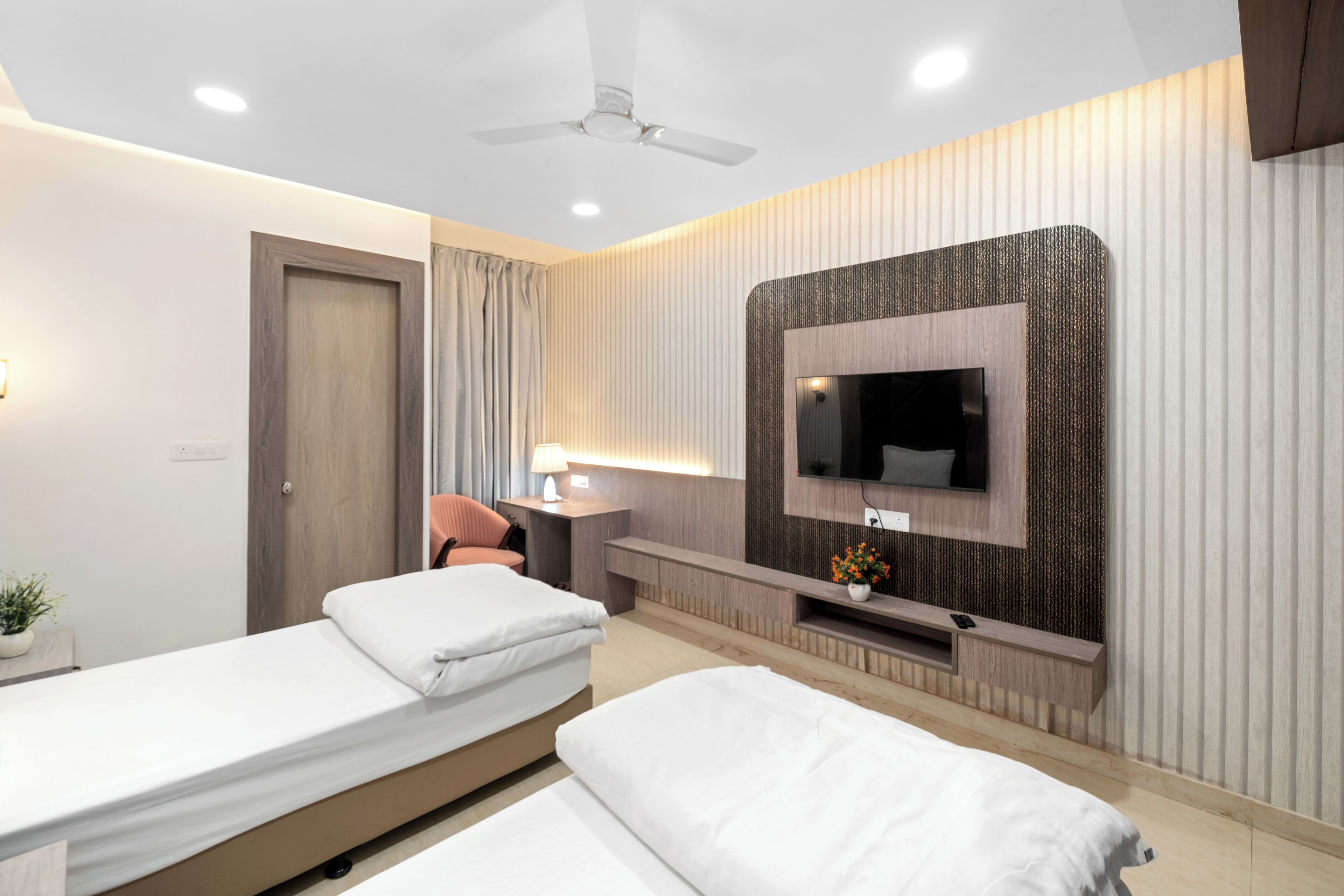 A modern hotel room with two beds, a TV mounted on the wall, a ceiling fan, and a small desk with a lamp.