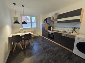 A modern kitchen with dark cabinetry, a built-in oven, and a dishwasher. There is a dining area with a table and chairs, and a window providing natural light. - Large modern vacation apartment (Kassel)