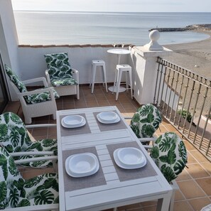 A terrace with a table set for two, white wicker furniture, and a view of the sea.