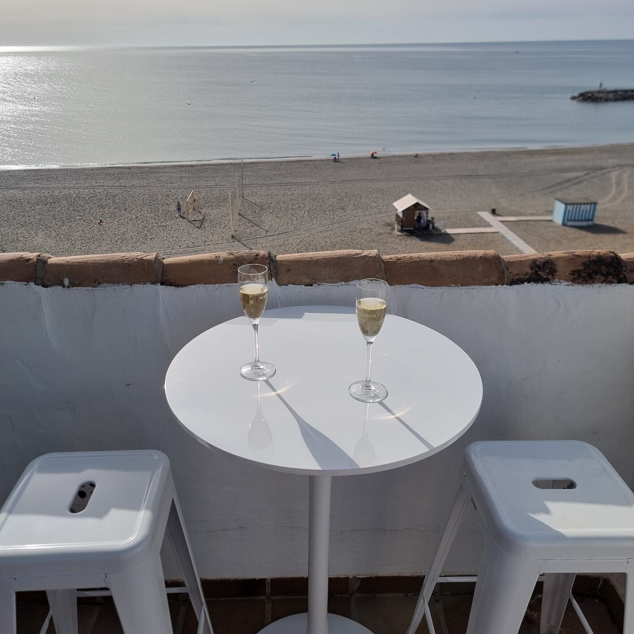 Un balcón con dos mesas blancas, cada una con una copa de vino blanco, con vista a una playa de arena y una estructura pequeña a lo lejos.