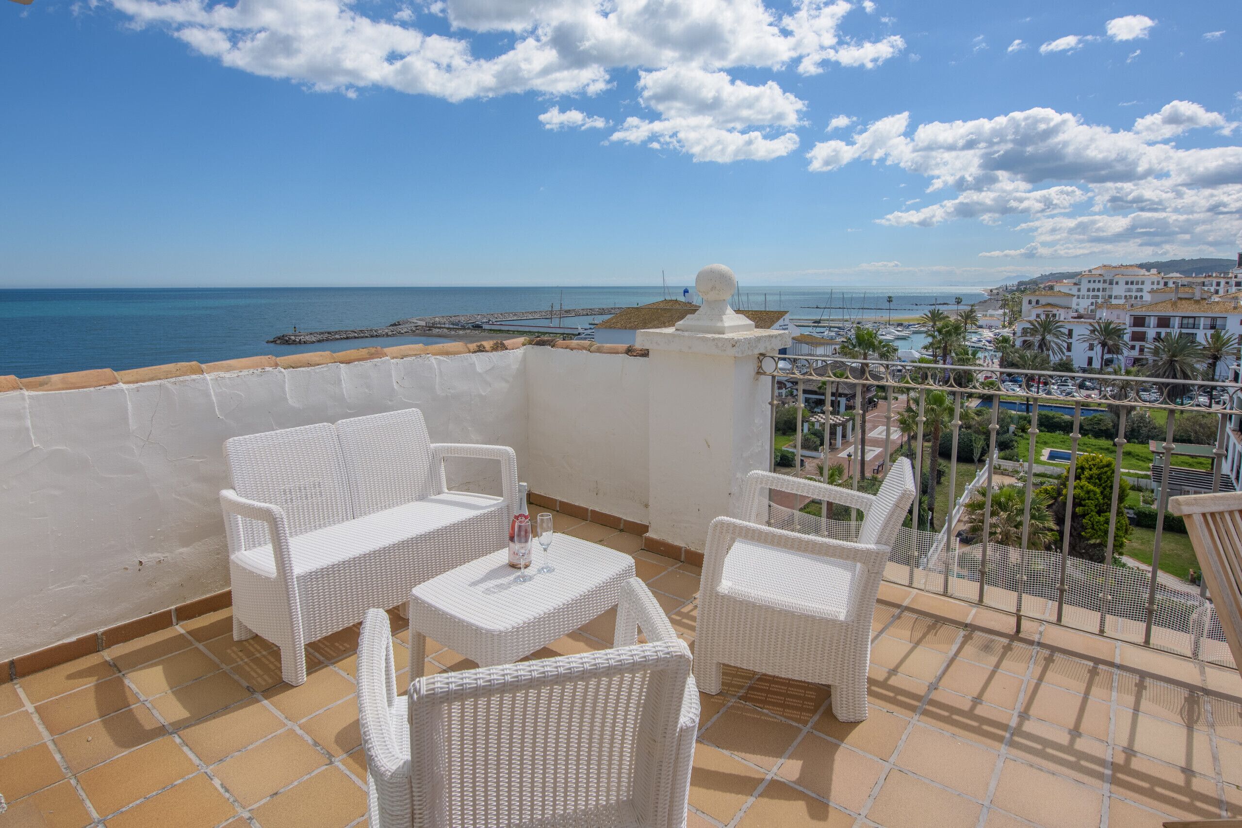 A balcony with white wicker furniture, a bottle of wine, and a view of the sea and buildings.