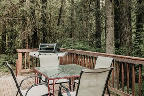 A deck with a grill, chairs, and a table surrounded by a forest.