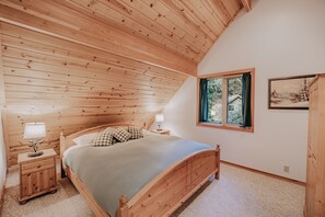 A bedroom with a wooden ceiling, a bed with a checkered pillow, a nightstand with a lamp, and a window with a view of trees.