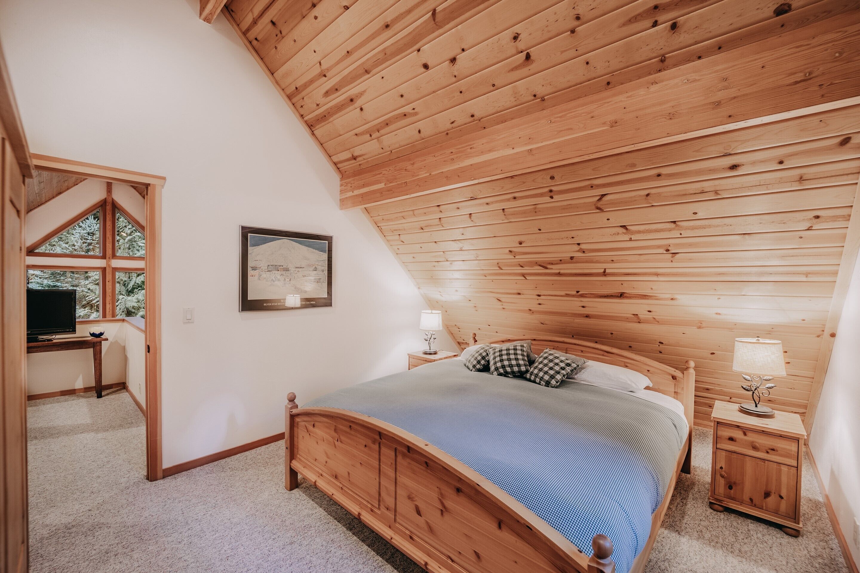 A bedroom with a sloped wooden ceiling, a bed with a blue bedspread, a nightstand with a lamp, and a desk with a television.