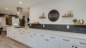 A modern kitchen with white cabinetry, a black backsplash, and a sign reading "Roots & Bourbon Bungalow." -  5-Bedroom Retreat with Cowboy Pool, Sauna and More – 10 Min to Downtown Austin (Austin)