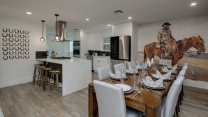 A modern kitchen with a central island, stainless steel appliances, and a dining area with a large mural. -  5-Bedroom Retreat with Cowboy Pool, Sauna and More – 10 Min to Downtown Austin (Austin)