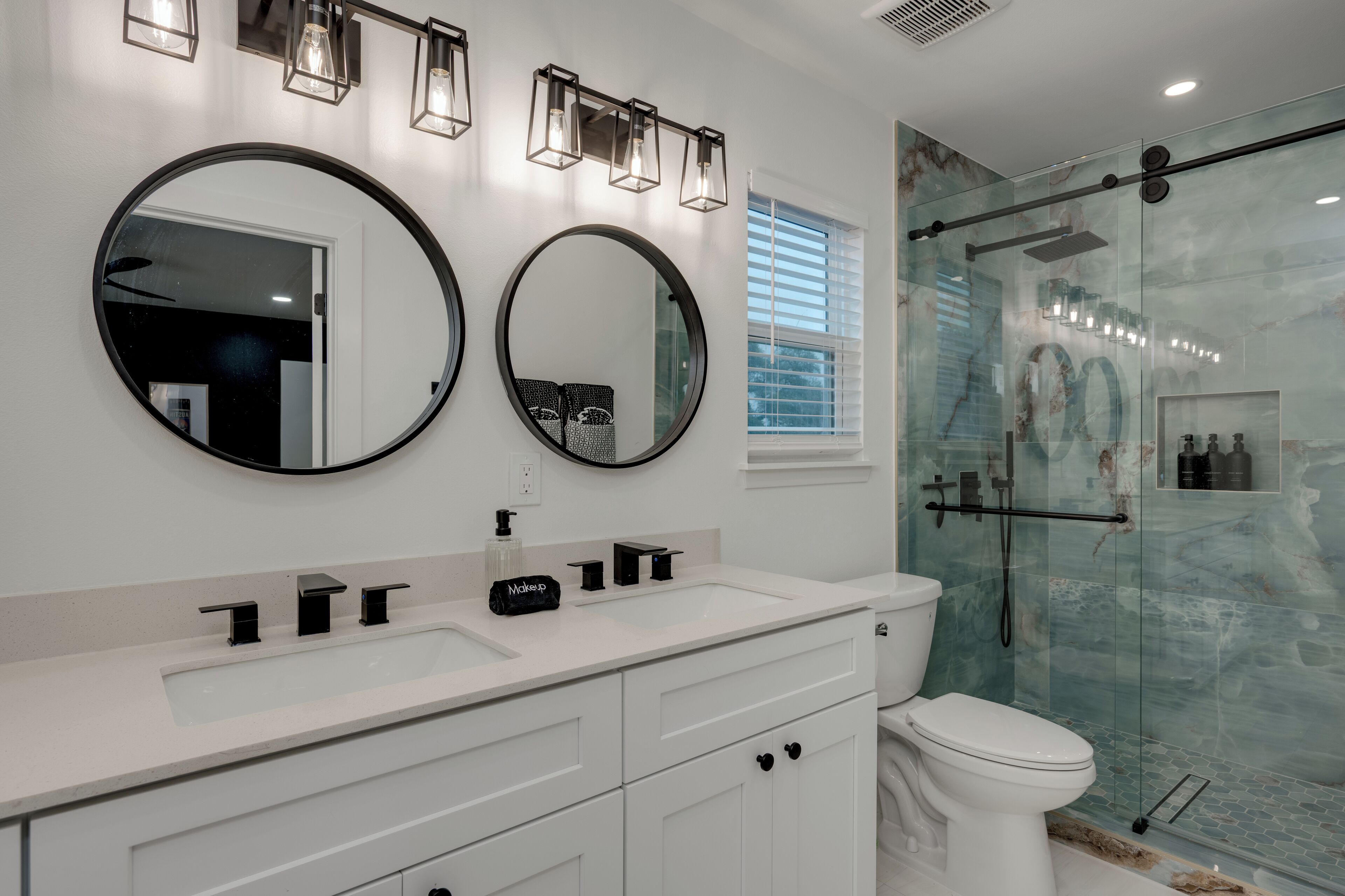 A modern bathroom with two round mirrors, a white sink, a toilet, and a glass-enclosed shower area.