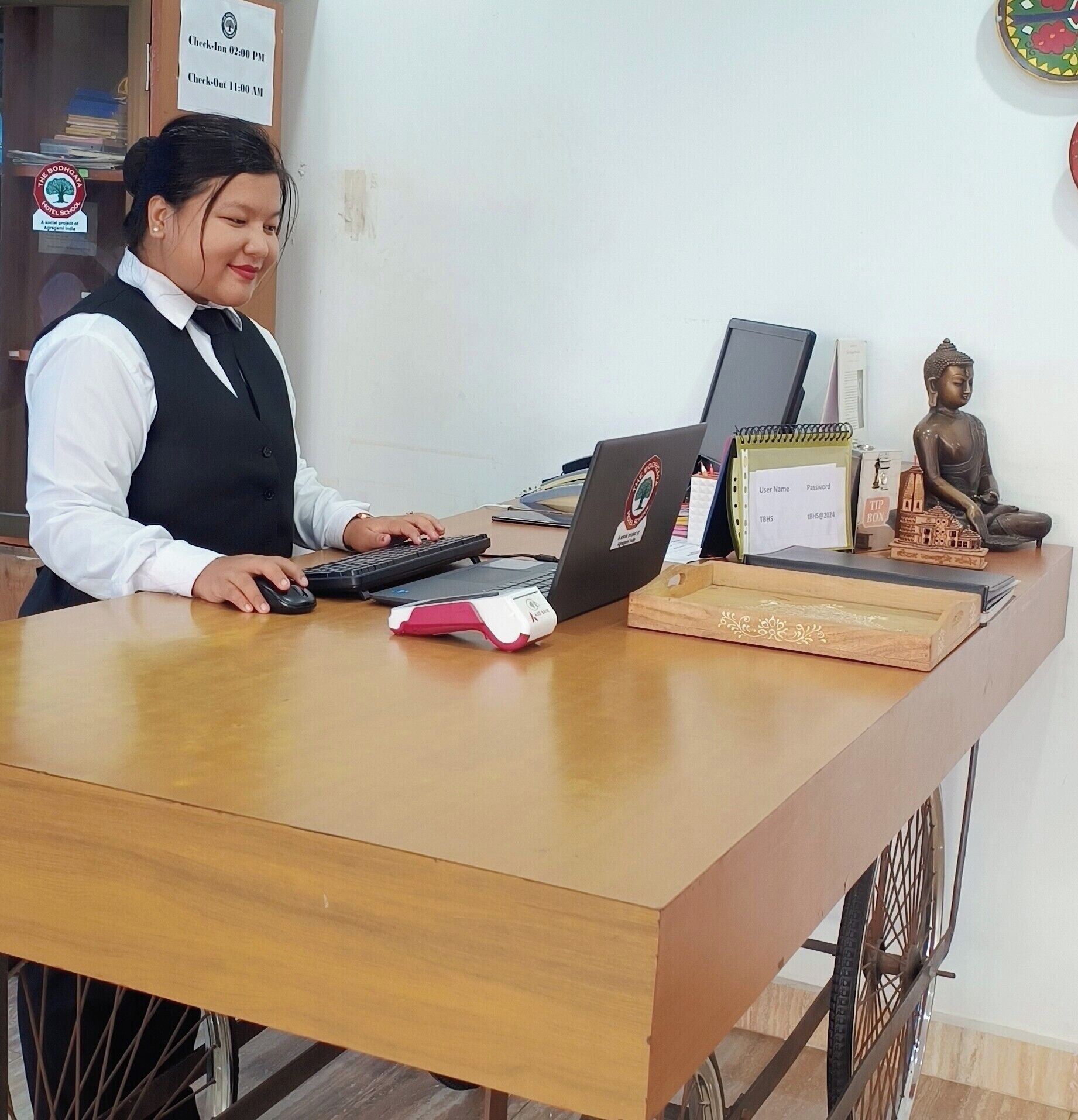 A reception desk with a laptop, a keyboard, a mouse, a check-in/out sign, and a Buddha statue.