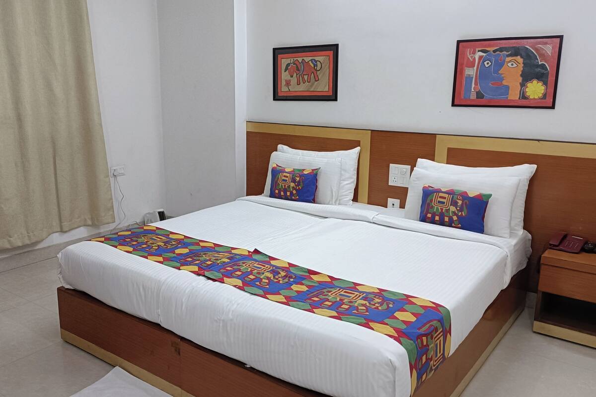 A hotel room with a large bed, white bedding, colorful throw pillows, and two framed artworks on the wall.