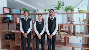Three individuals in formal attire standing in a well-organized room with wooden shelves containing books, decorative items, and potted plants. - The Bodhgaya Hotel School (Bodh Gaya)