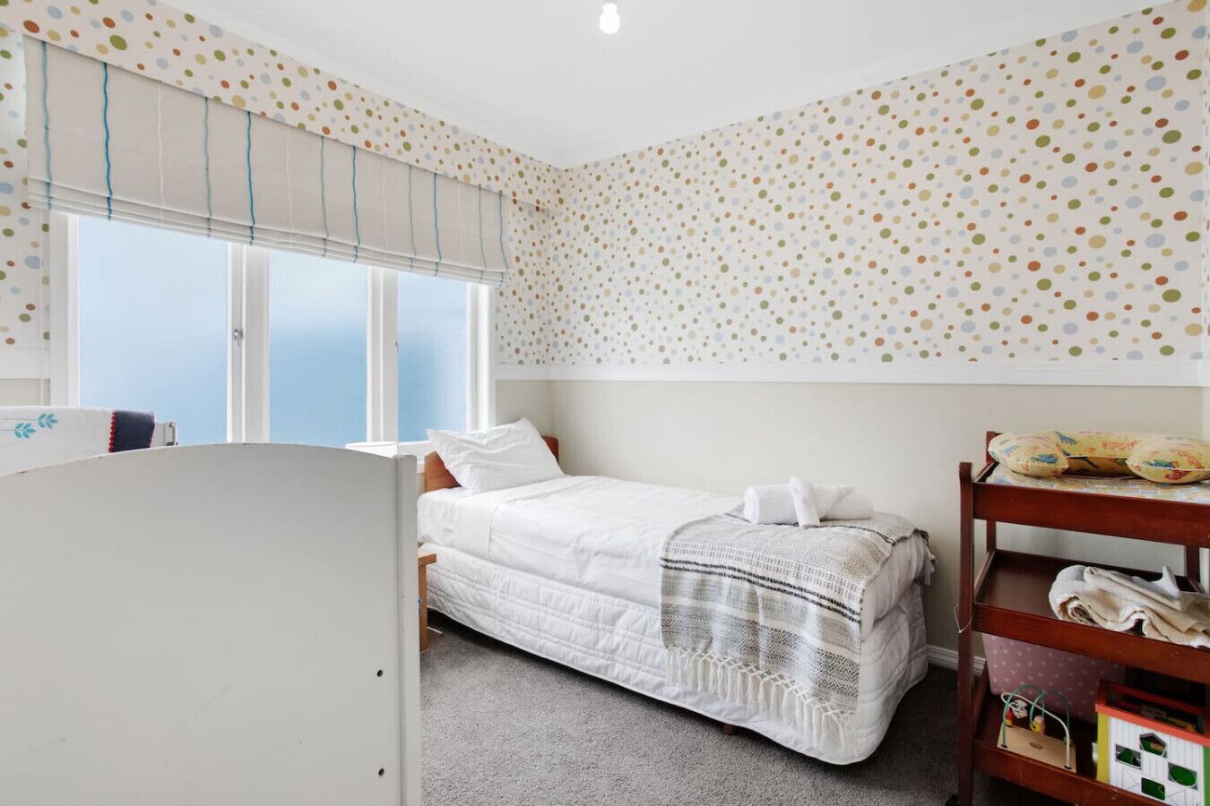 A bedroom with a single bed, a window with blinds, a wooden shelf with toys, and a polka dot wall.