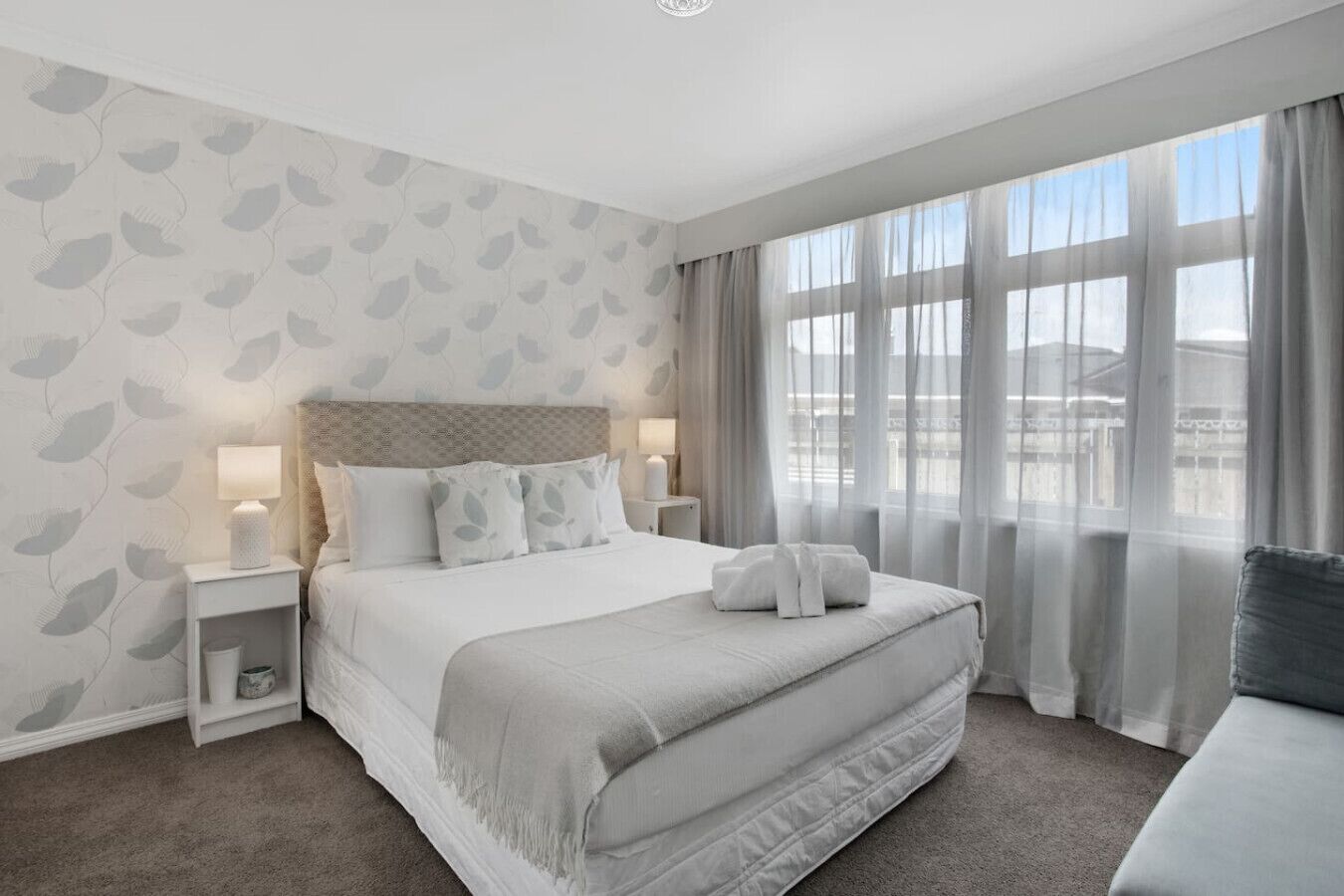 Une chambre d’hôtel moderne avec un grand lit, deux tables de chevet avec des lampes, une vue sur la ville à travers de fines rideaux, et un canapé gris.