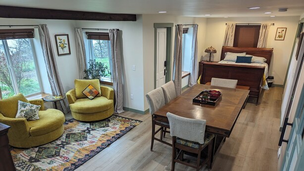 A spacious living area with a wooden dining table, two yellow armchairs, a patterned rug, and a view of the outdoors through large windows. - Charming studio. Close to highway and ferries. (Bremerton)