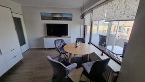 Una sala de estar moderna con una mesa redonda de madera, sillas de cuero, un televisor de pantalla plana y un ventanal con vistas a edificios.
