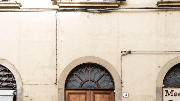 Une porte en bois à deux battants, ornée d’une grille métallique, est encadrée dans un bâtiment de couleur beige, devant lequel se trouve une rue pavée de pierres.