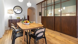 Un coin repas avec une table en bois, des chaises noires et un saladier rempli de fruits. On y trouve des luminaires suspendus et une grande fenêtre avec des cadres en bois.