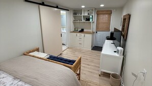 Een slaapkamer met een bed, een klein bureau, een stoel en een kitchenette op de achtergrond.