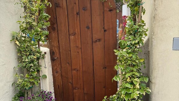 Een houten poort met een wijnrank erboven, beklemd tussen twee zwarte potten met paarse bloemen.