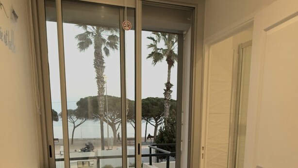 A balcony with a glass door, two white chairs, a white table, and a view of palm trees and a park.