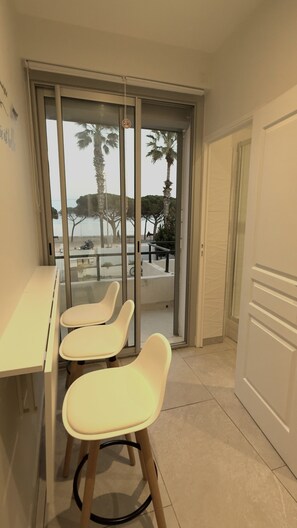 A balcony with a glass door, two white chairs, a white table, and a view of palm trees and a park. - Seaside apartment (La Ciotat)