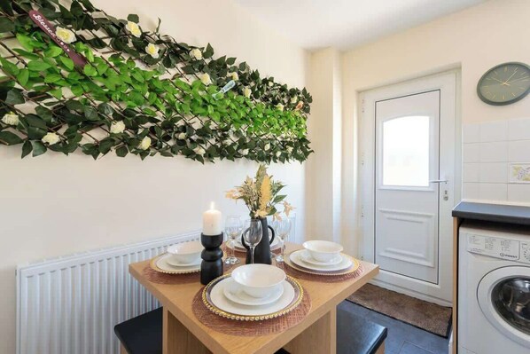 A dining area with a table set for four, a decorative wall hanging, and a washing machine in the background.